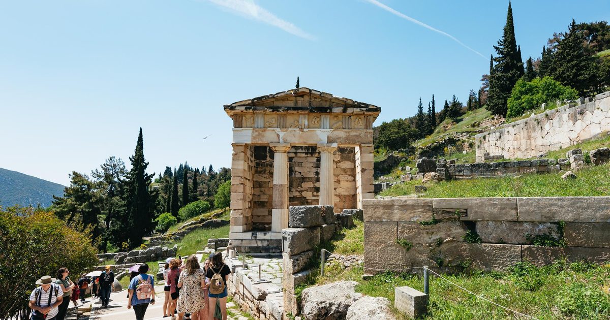 Visitors on the Sacred Way at Delphi passing the rebuilt Treasury of the Athenians, with cypress trees on the slope behind