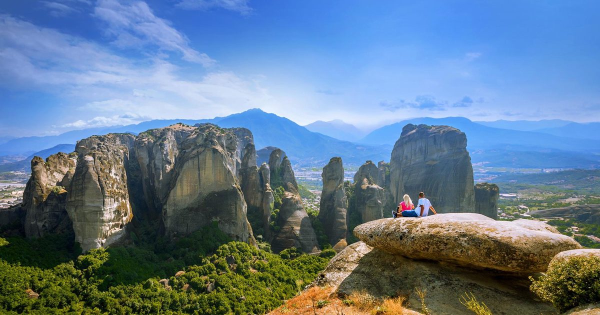 Two visitors sitting on a rock ledge above the sandstone pillars of Meteora, with the plain of Thessaly and mountains beyond